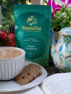 Mamma Chai Assam Chai Blend package with a cup of tea, cookies, and a teapot on a table.