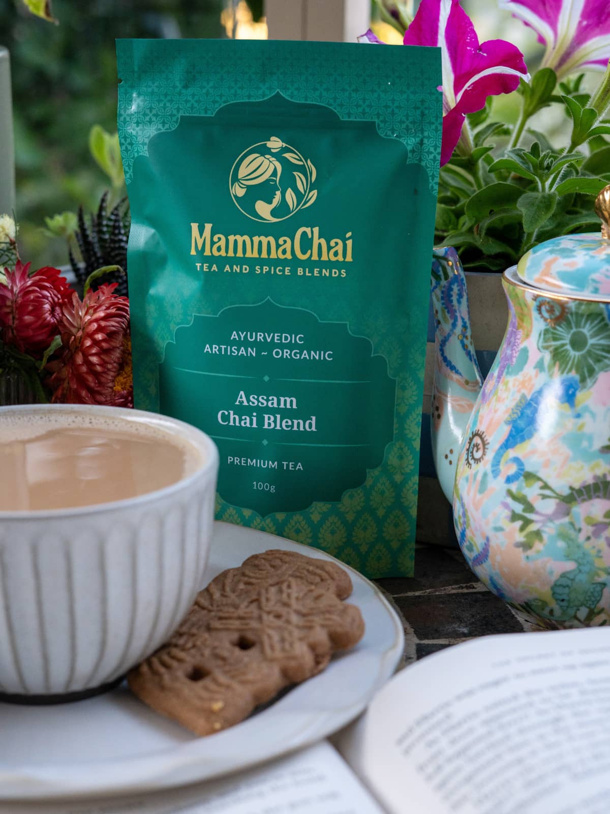 Mamma Chai Assam Chai Blend package with a cup of tea, cookies, and a teapot on a table.